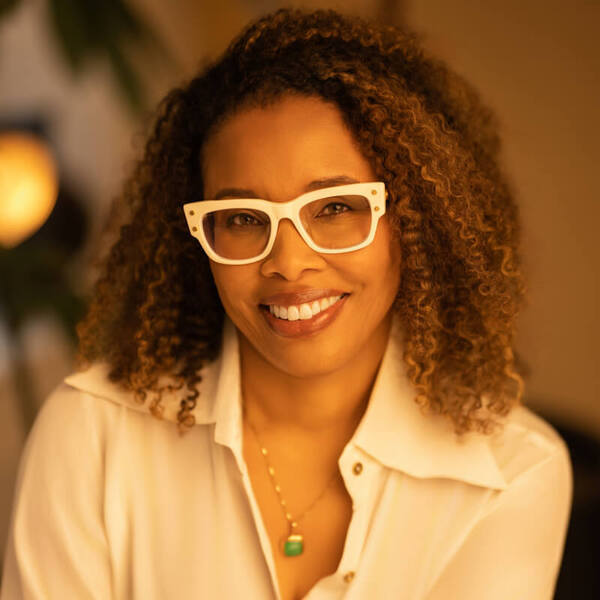 A woman with curly brown hair and white-framed glasses smiles, wearing a white shirt and a pendant.