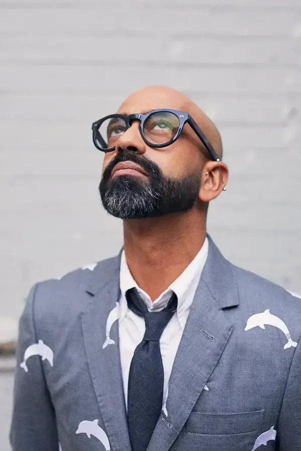 A person with no hair and light brown skin wearing a grey/blue blazer with white dophins on it is staring up at the sky through black glasses